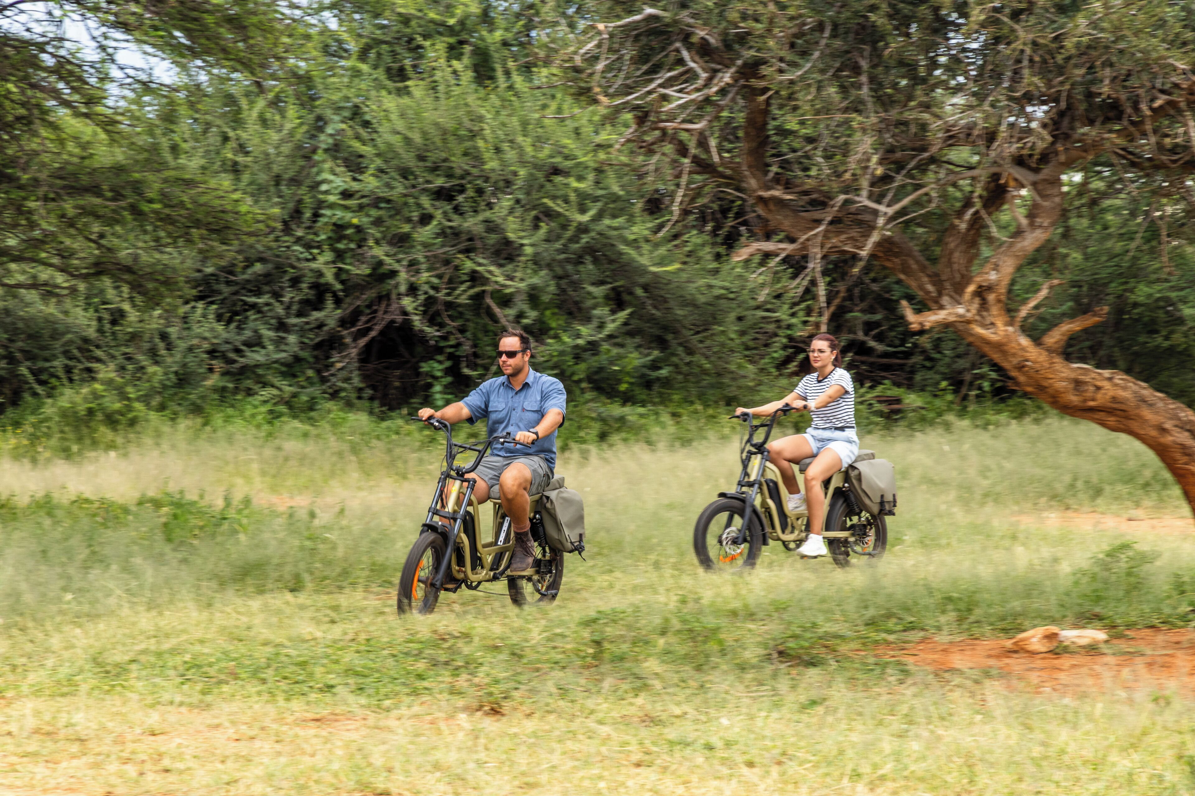 Two people riding electric bikes in a grassy area with trees in the background.