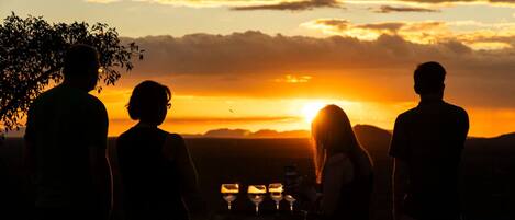 A group of people silhouetted against a sunset, standing near a table with glasses and bottles.