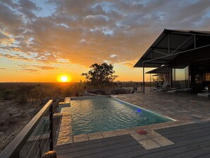 Una piscina al aire libre con vistas al atardecer, rodeada de sillones de descanso y un porche cubierto.