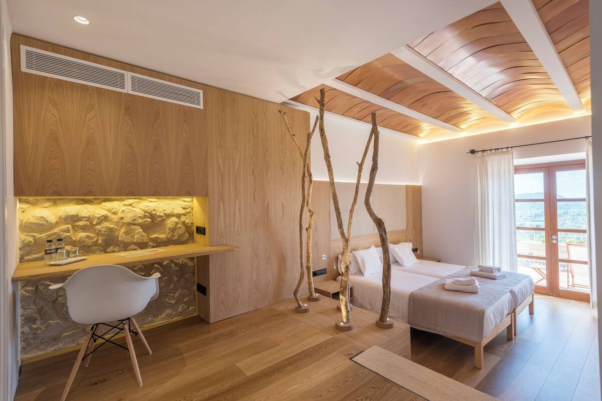 Une chambre d’hôtel moderne avec un plafond en bois, un mur décoratif en pierre, un bureau avec une chaise, un lit avec deux oreillers et un balcon avec vue.