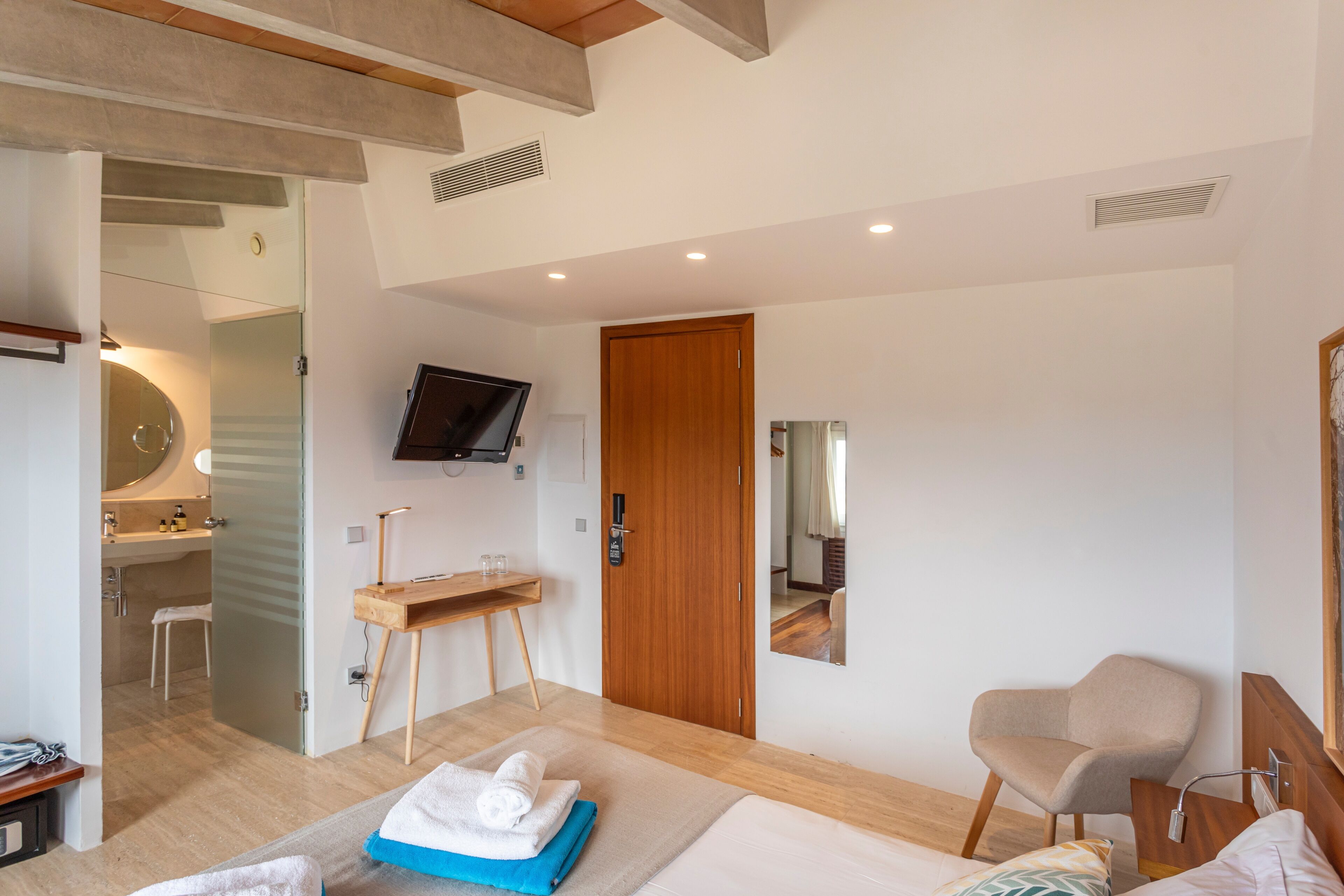 A modern hotel room with a bed, a wooden desk, a chair, a television, and a bathroom visible through an open door.