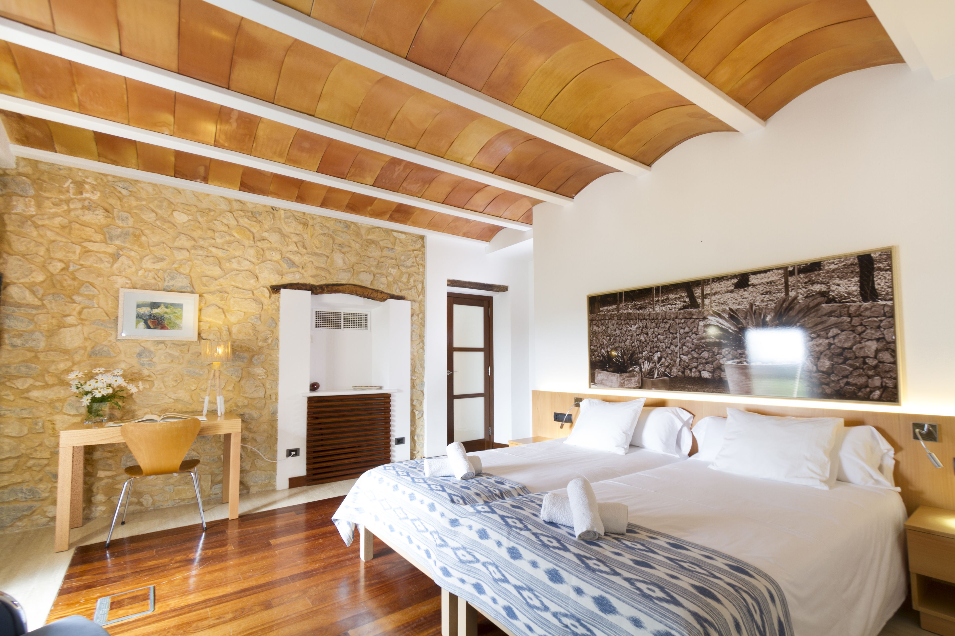 A bedroom with a large bed, a desk with a chair, a stone wall, and a wooden ceiling.