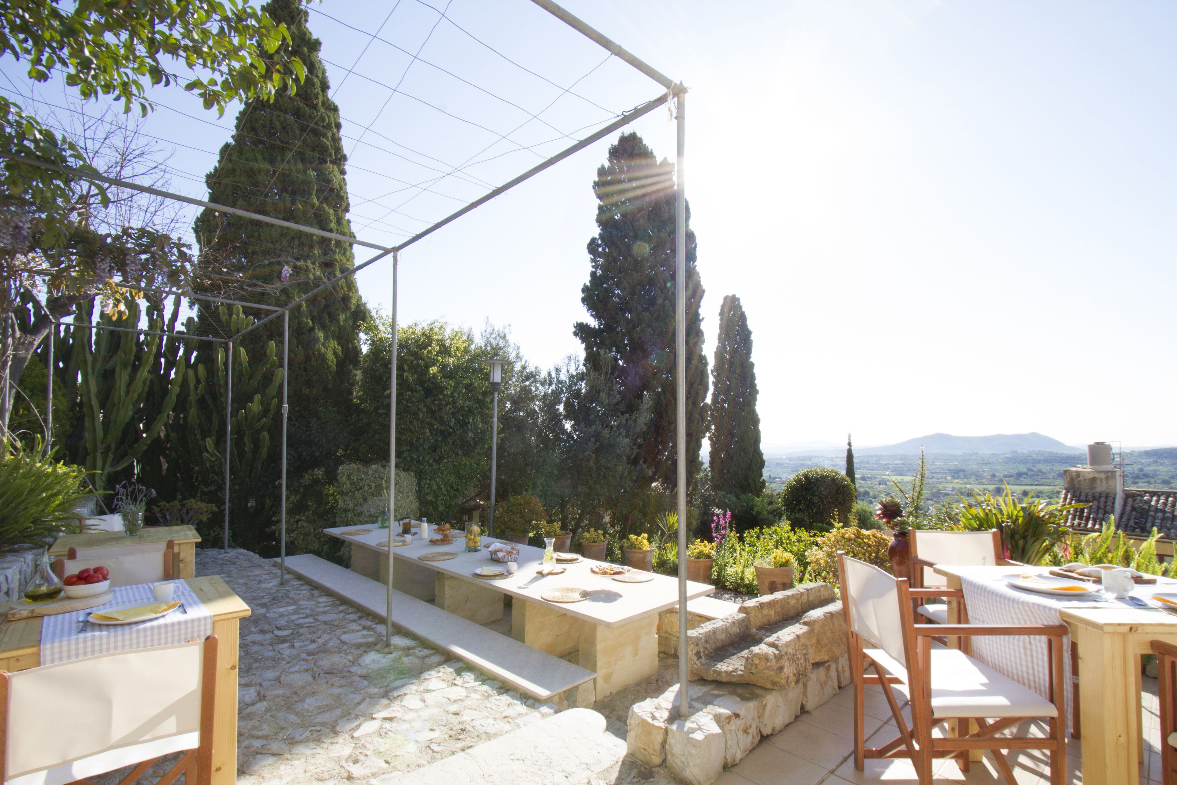 An outdoor dining area with stone flooring, wooden tables and chairs, and a variety of potted plants.