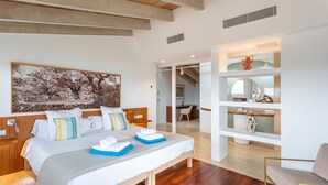 Habitación de hotel moderna con una cama grande, cabecero de madera y un cuadro con árboles. Cuenta con un área de descanso con una silla y una mesita.