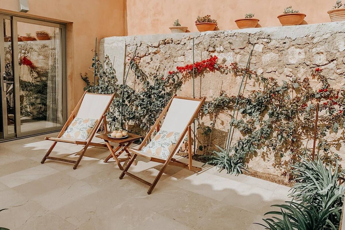 Une terrasse avec deux fauteuils de jardin en bois, un mur de pierre orné de plantes grimpantes et une porte vitrée.