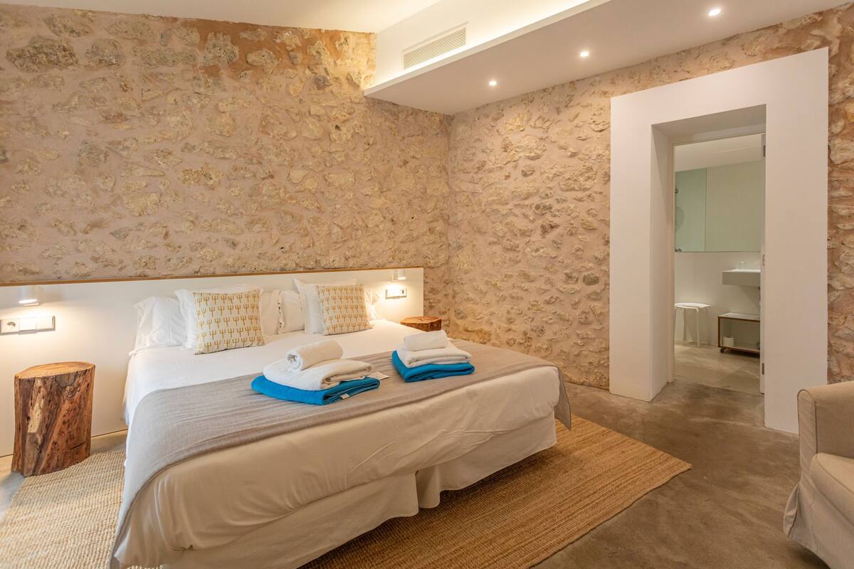 Une chambre avec un mur en pierre, un grand lit recouvert de draps blancs, une table de chevet en bois et un petit tabouret en bois.