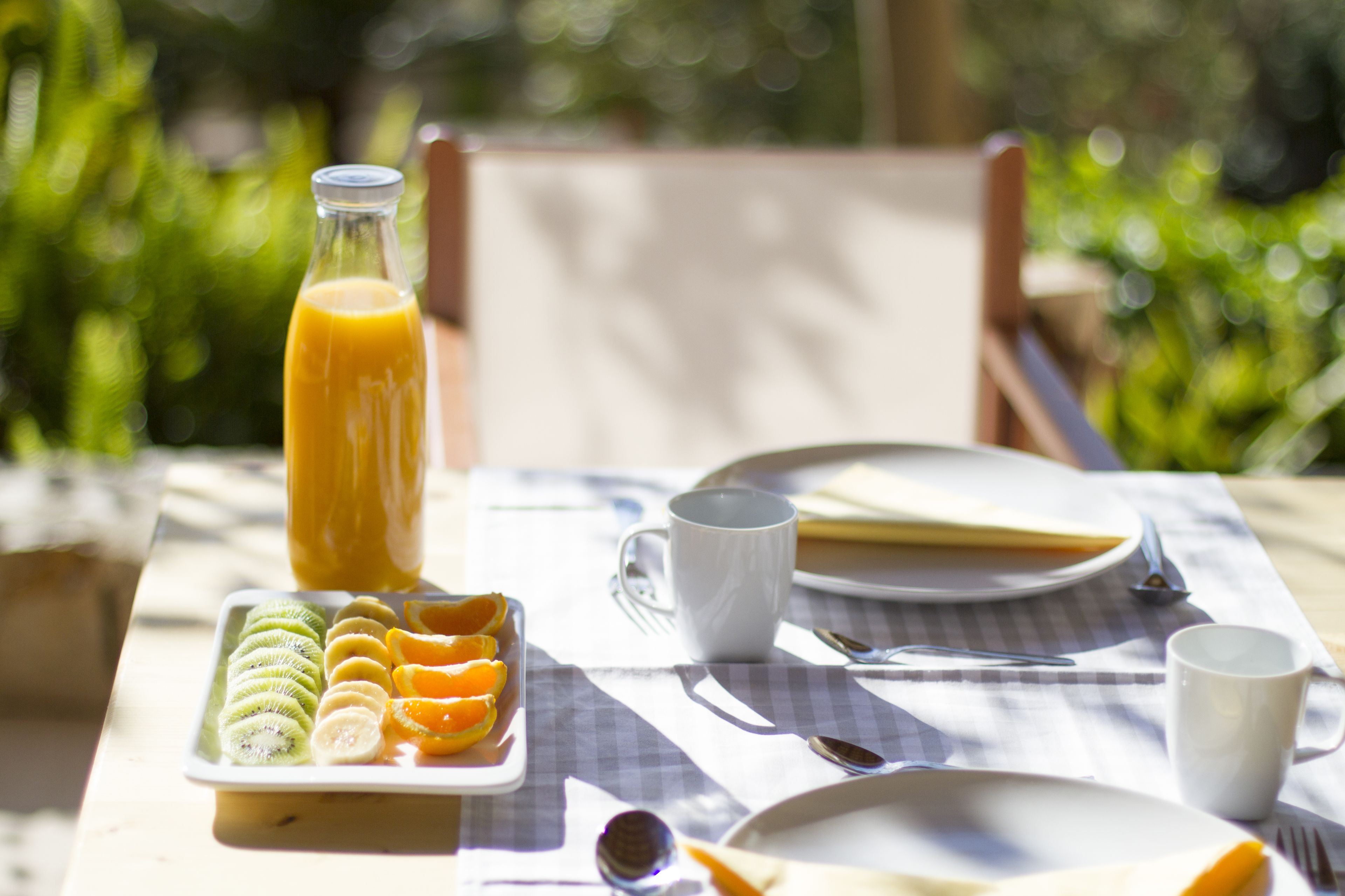 A breakfast table with a bottle of orange juice, a plate of sliced fruits, a white mug, and a checkered tablecloth.