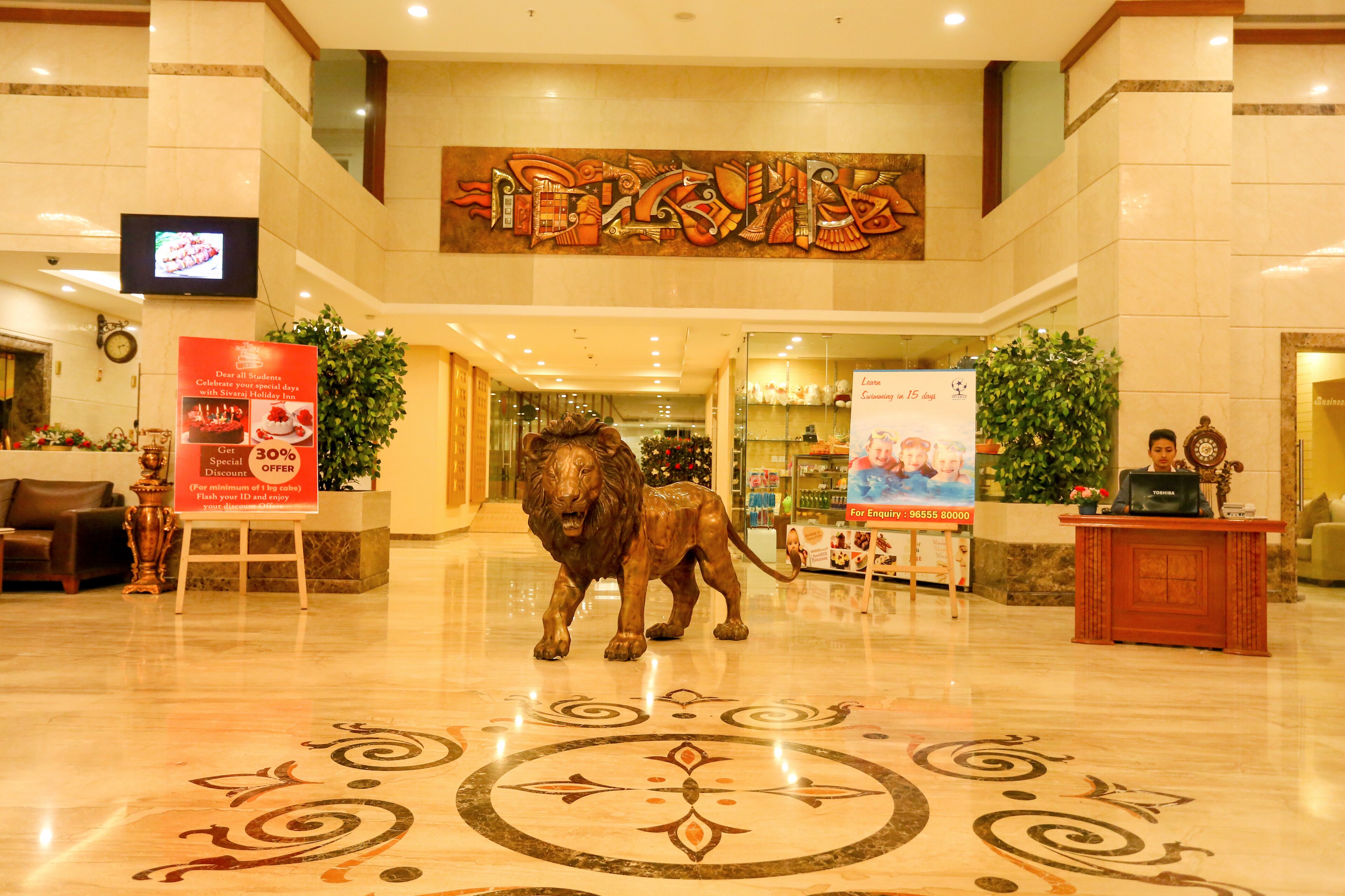 A lobby with a large lion statue, a reception desk, a clock, and a TV screen.