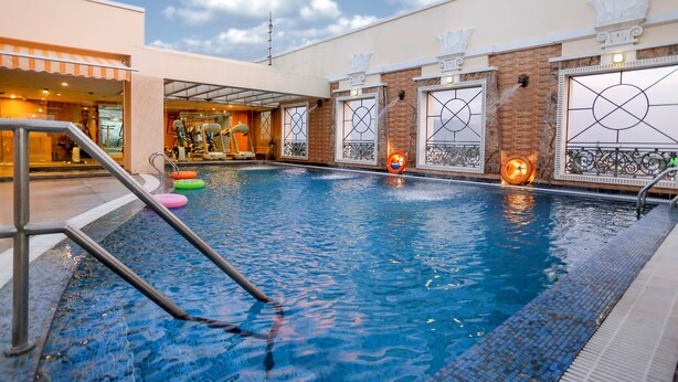 Una piscina interior con agua cristalina, rodeada por un solado de azulejos y un edificio con amplias ventanas y barandillas metálicas decorativas.