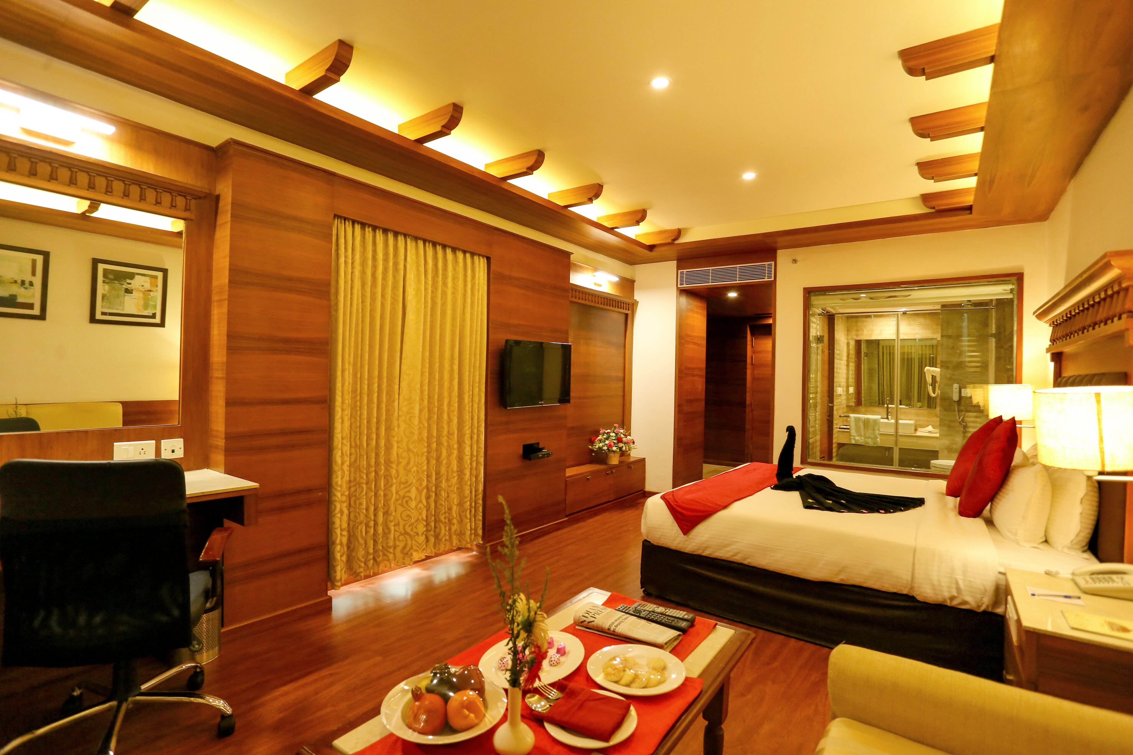 A hotel room with a bed, a desk, a chair, a dining table with plates and a vase, and a television on the wall.