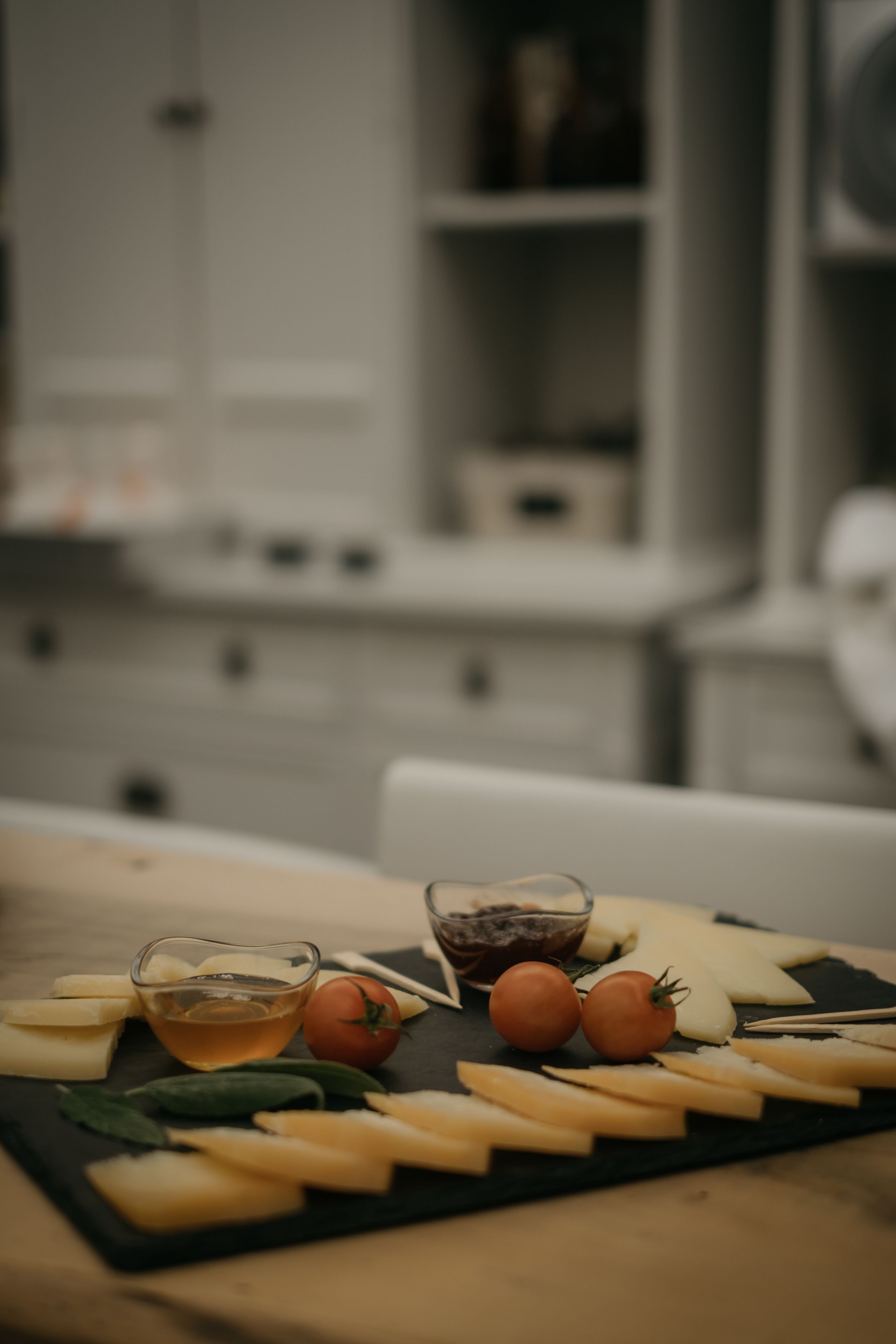 A cheese board with various cheeses, tomatoes, and a small bowl of sauce on a dark slate board.