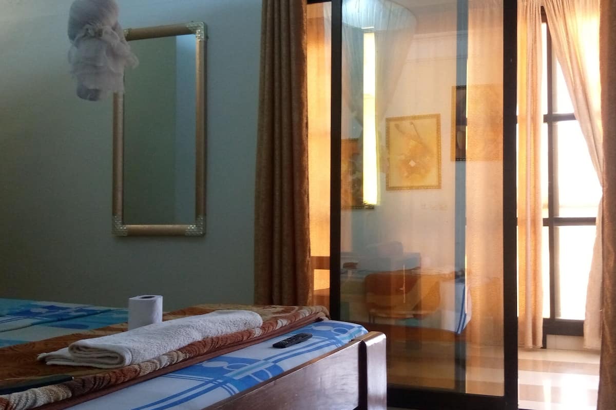 A hotel room with a bed, a mirror, a towel, a remote control, and a view of the outside through a glass door.