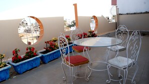 A rooftop terrace with a round glass table and white metal chairs, surrounded by blue flower planters with red and yellow flowers. - Residence Les Calanques (Dakar)