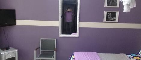 Un dormitorio con una cama, una silla, un espejo y un televisor sobre una mesita de noche.