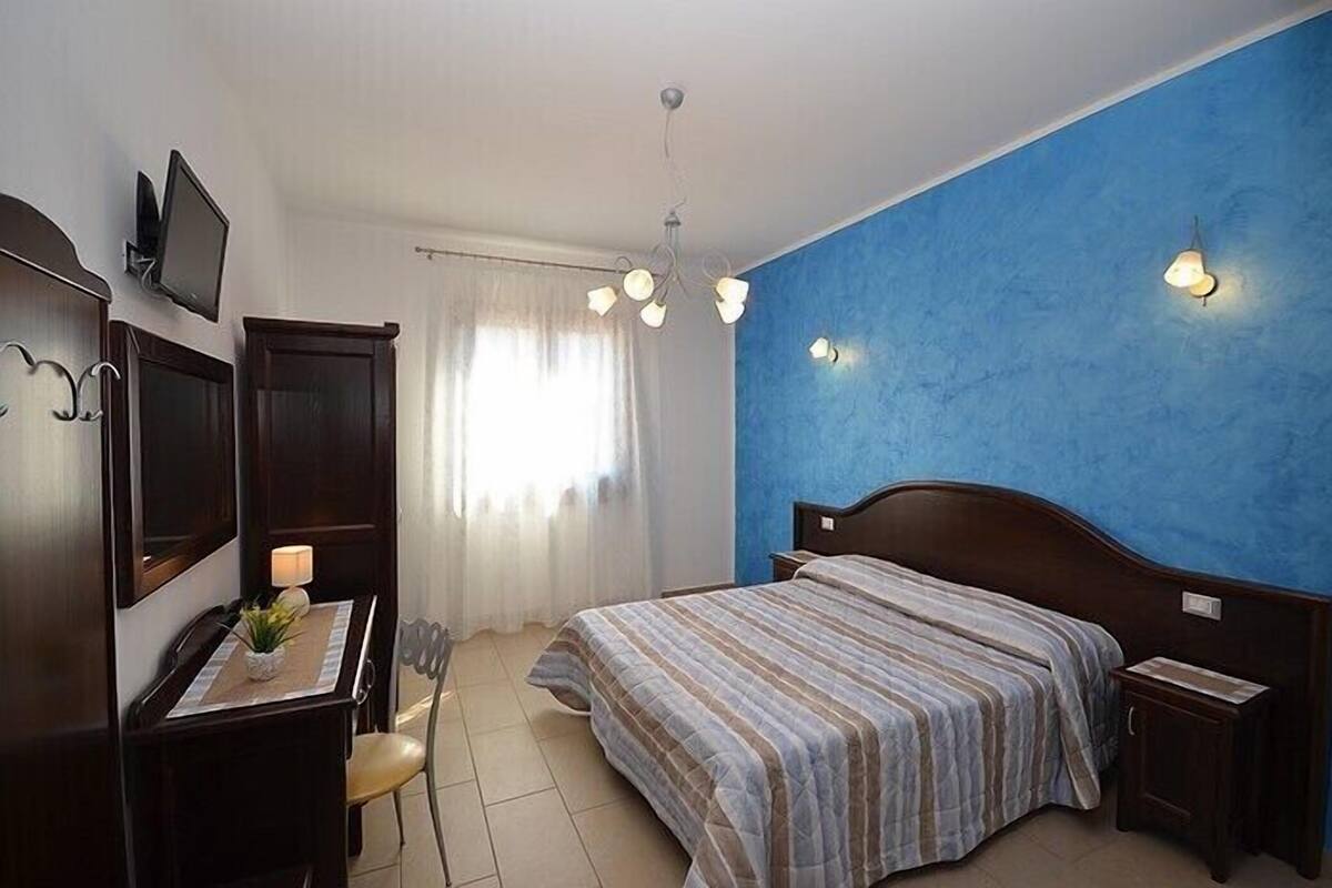A bedroom with a bed, a desk with a lamp, a chair, a TV, and a blue accent wall.