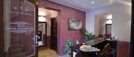 A hotel reception area with a glass door labeled "Baia Santa Margherita" and a desk with a plant and papers.