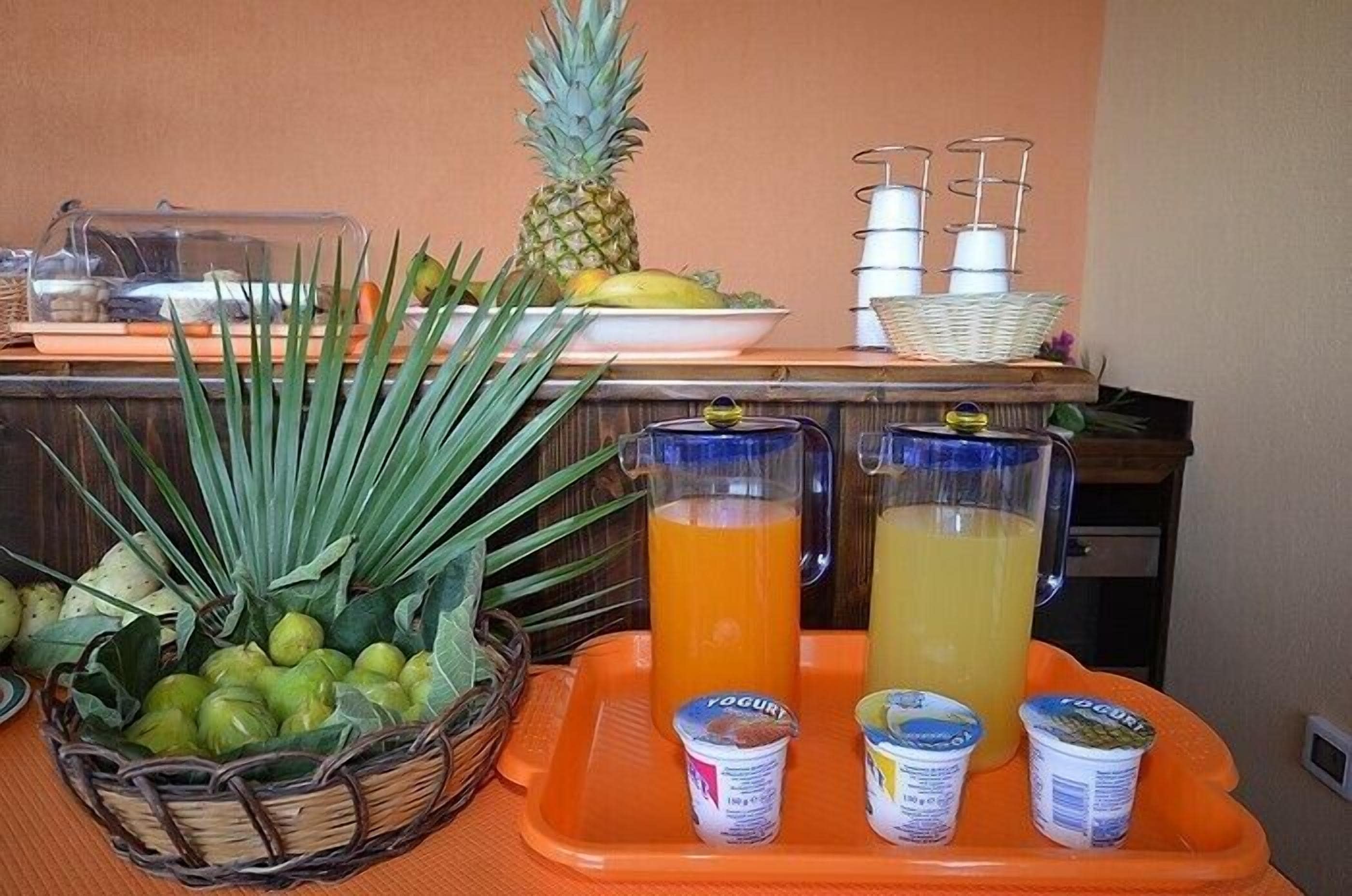 A buffet setup with a pineapple, a basket of fruits, and two pitchers of orange juice and yogurt.