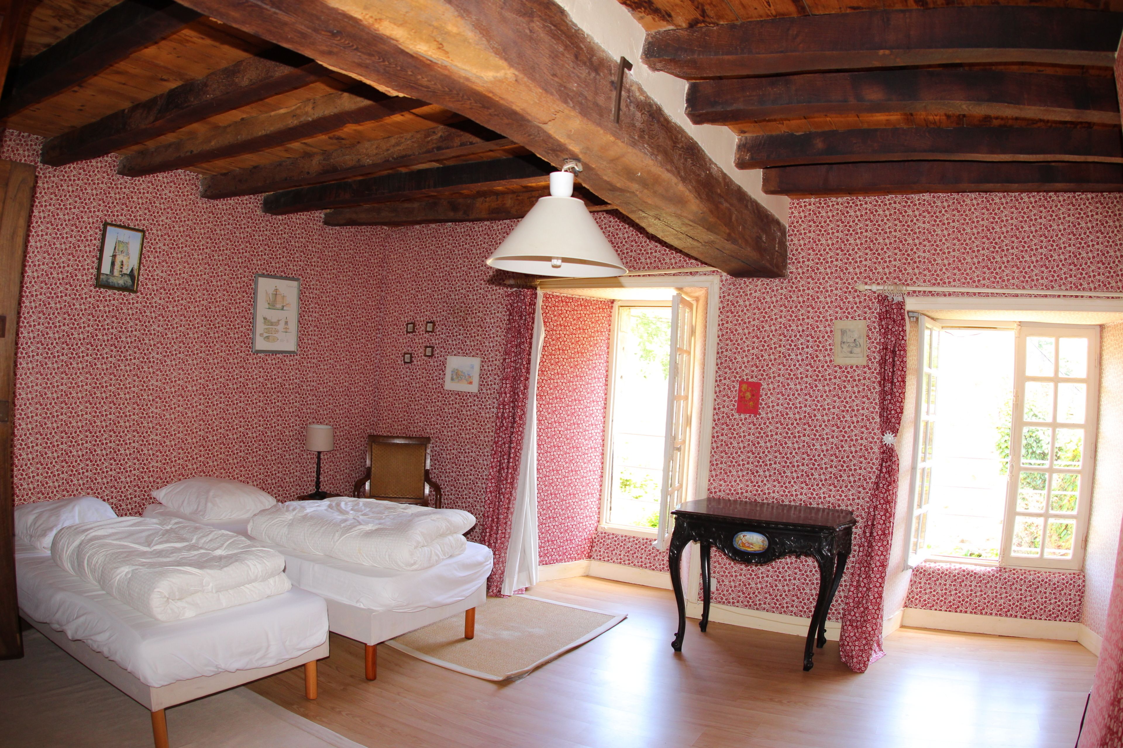 Una habitación con dos camas, vigas de madera en el techo, una mesa y un ventanal con cortinas.