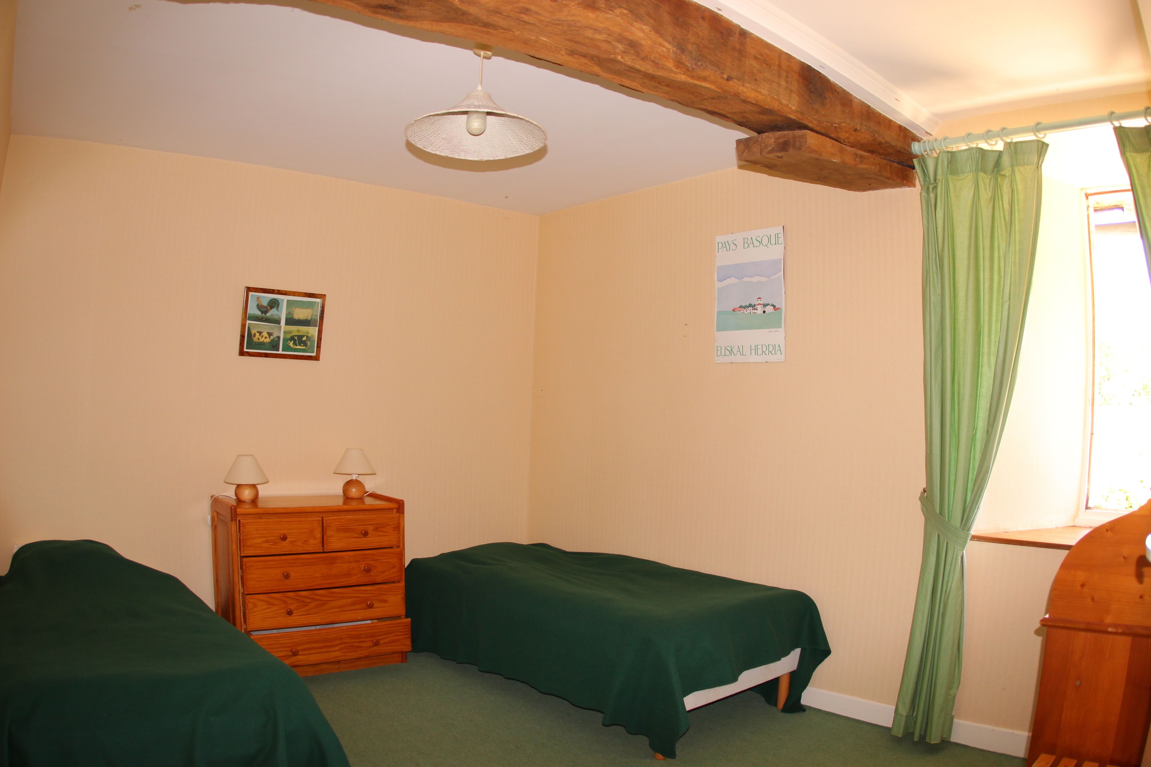 Een kamer met twee enkele bedden, een houten dressoir, een klein fotolijstje en een raam met groene gordijnen.