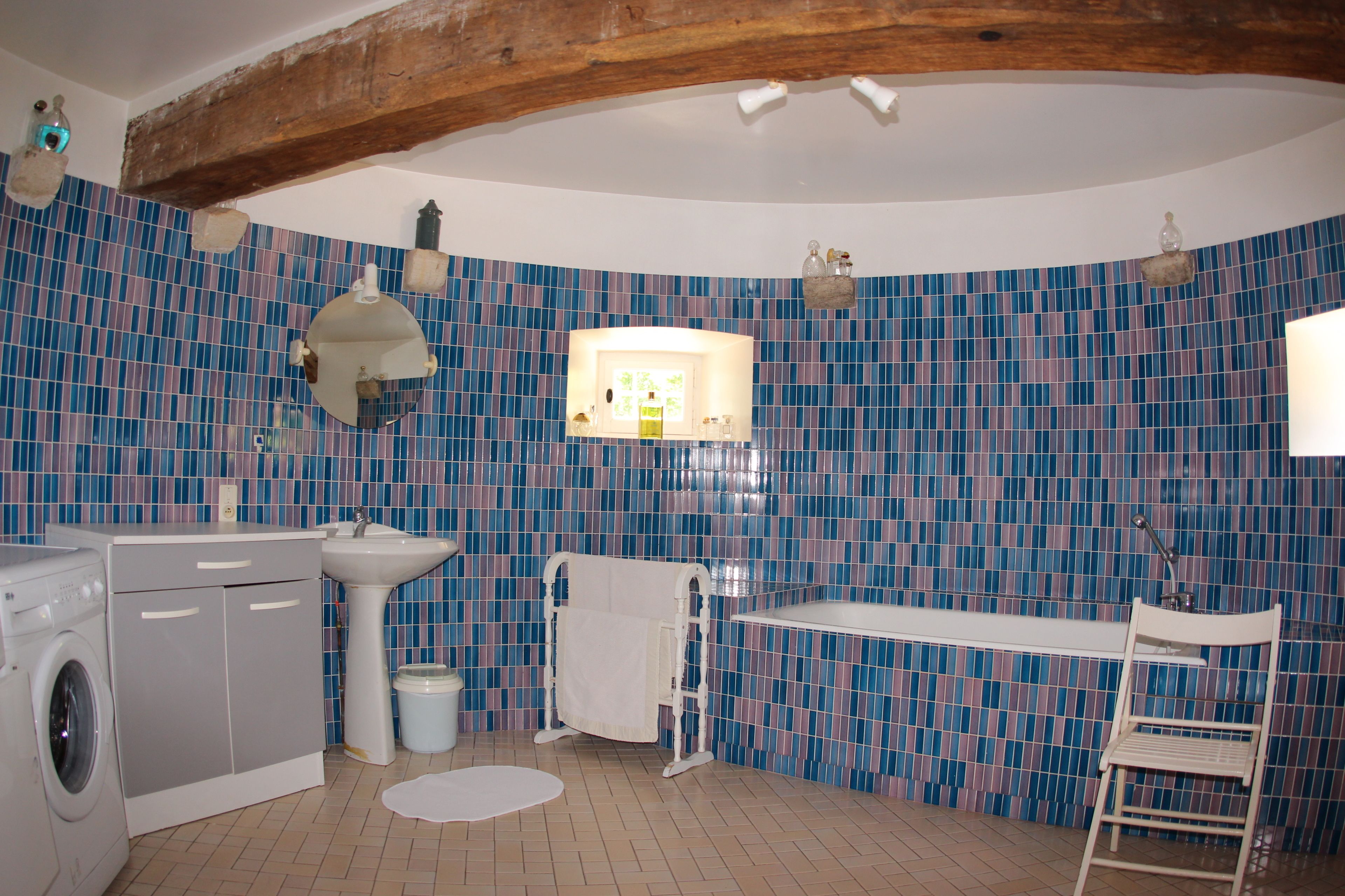 A bathroom with a tiled wall, a sink, a mirror, a towel rack, a shower, and a washing machine.