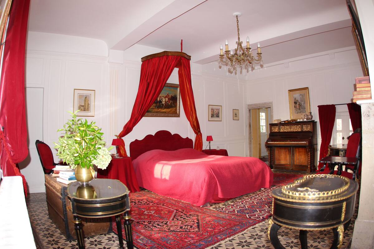 Una habitación con cama con dosel, una araña, un dosel rojo, un armario de madera y una alfombra estampada.