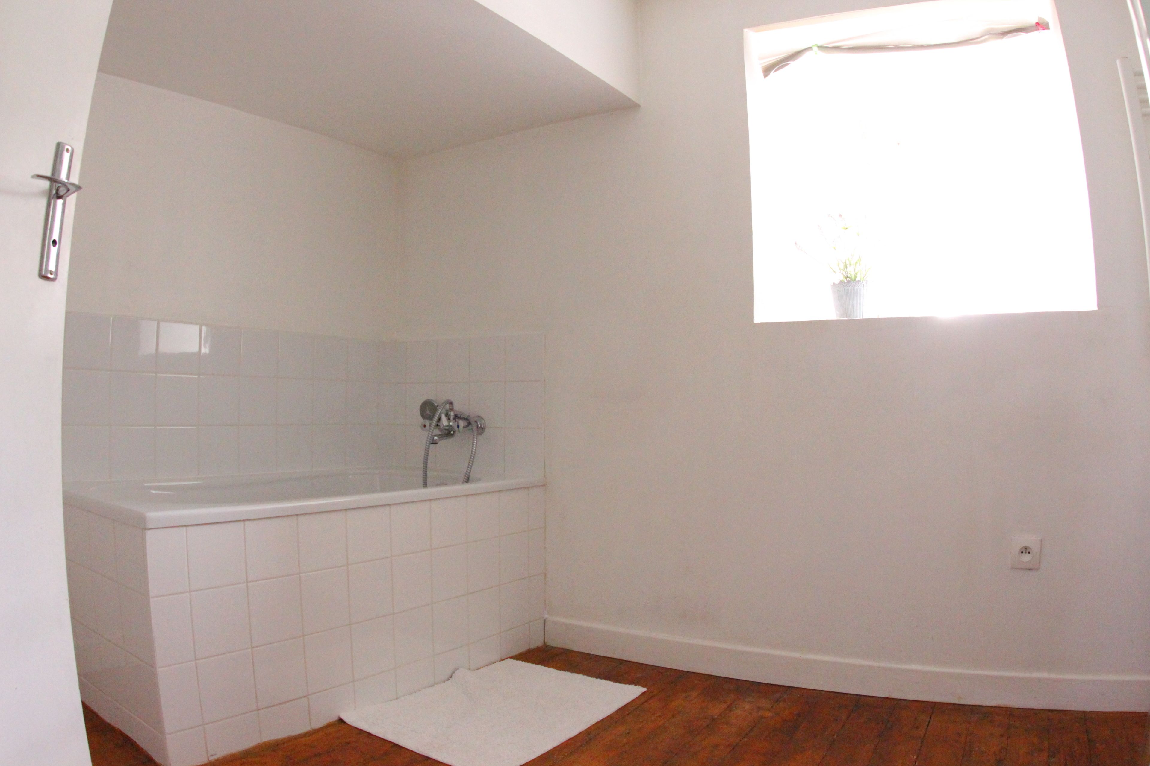 Een badkamer met een witte badkuip, een klein raam met een plant, en een houten vloer.
