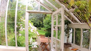 Une véranda avec une table et des chaises en bois, entourée d’une végétation luxuriante et de fleurs.