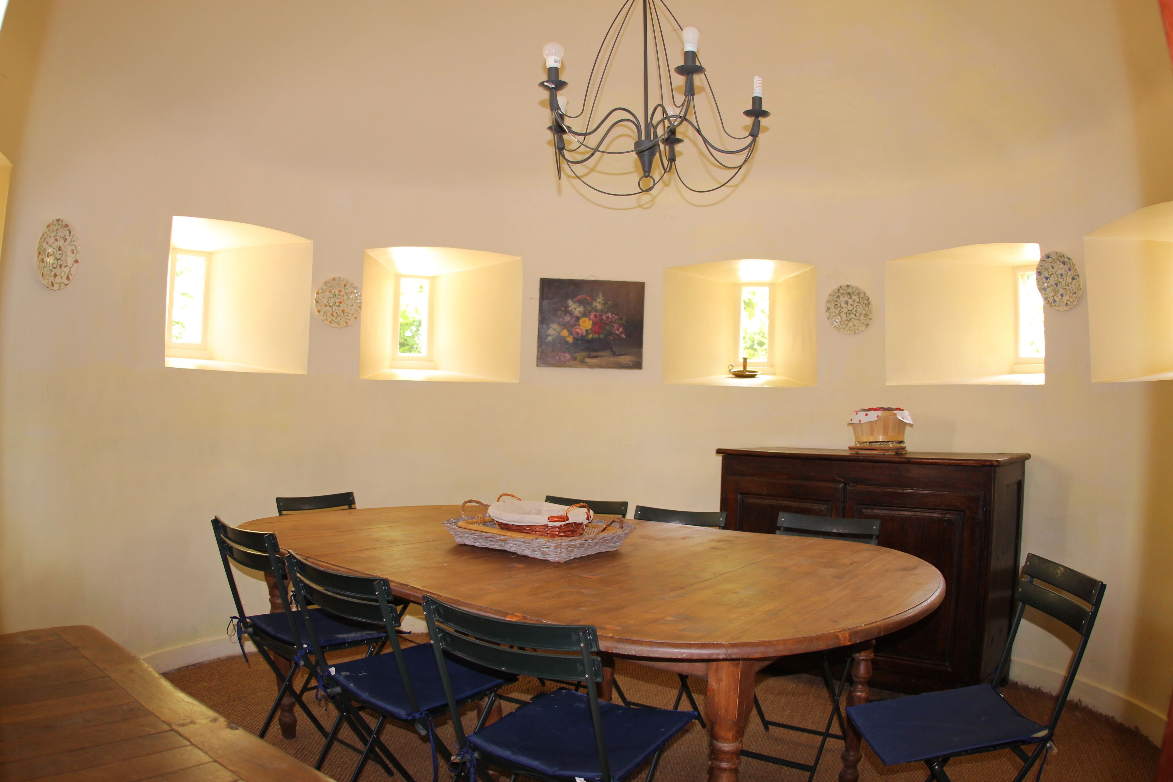 Un comedor con una mesa redonda de madera, seis sillas, una lámpara de araña, lámparas de pared y un cuadro.