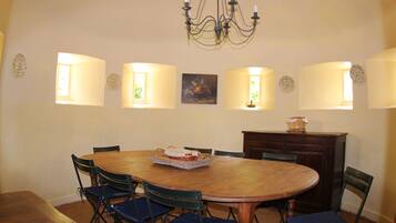 Un comedor con una mesa redonda de madera, seis sillas, una lámpara de araña, lámparas de pared y un cuadro.