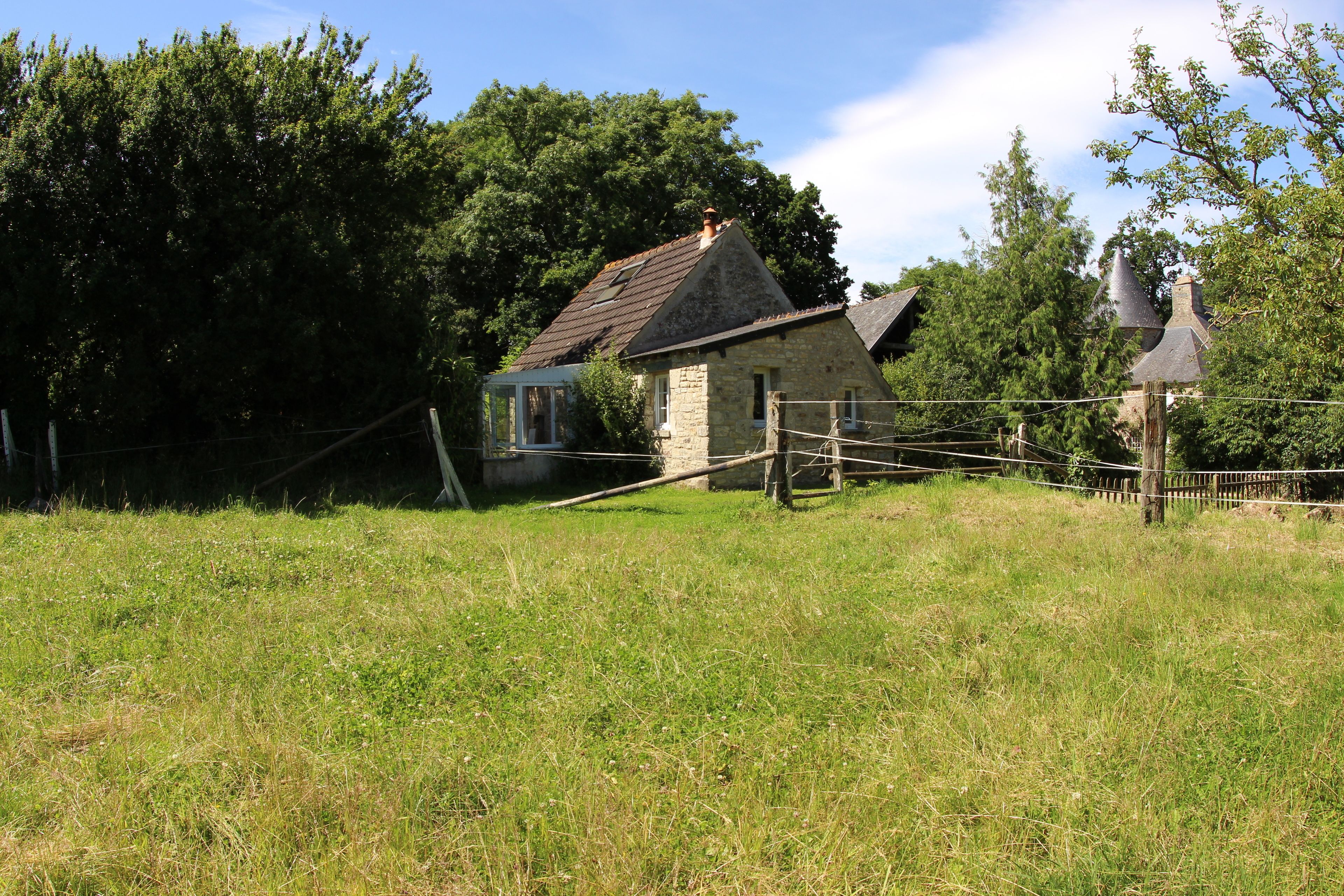 Ein Steinhaus mit geneigtem Dach, umgeben von einem Holzzaun und üppiger Bepflanzung.
