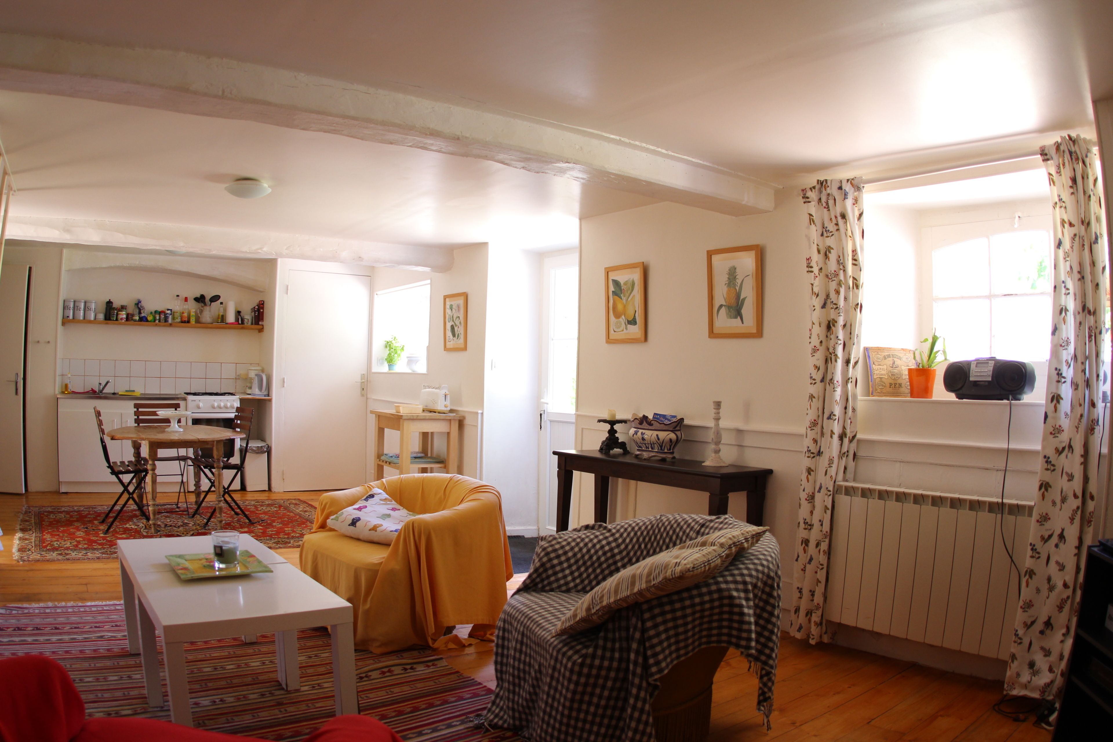 Sala de estar con sofá, una mesa, una silla, una zona de cocina con estufa y alacenas, y una ventana con cortinas.