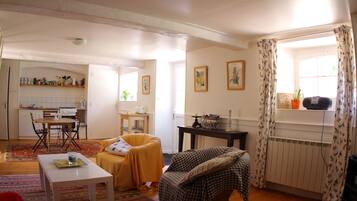 Sala de estar con sofá, una mesa, una silla, una zona de cocina con estufa y alacenas, y una ventana con cortinas.