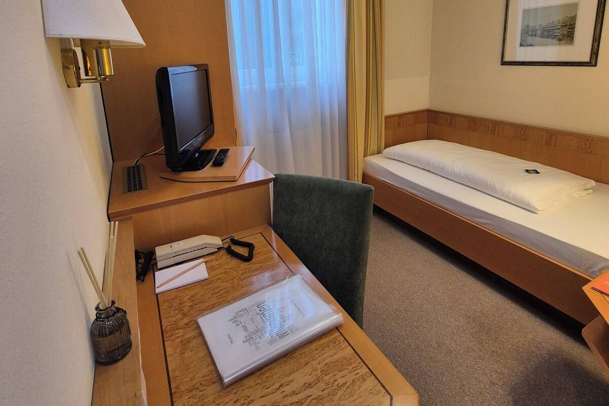 A hotel room with a wooden desk, a television, a bed, and a window with curtains.