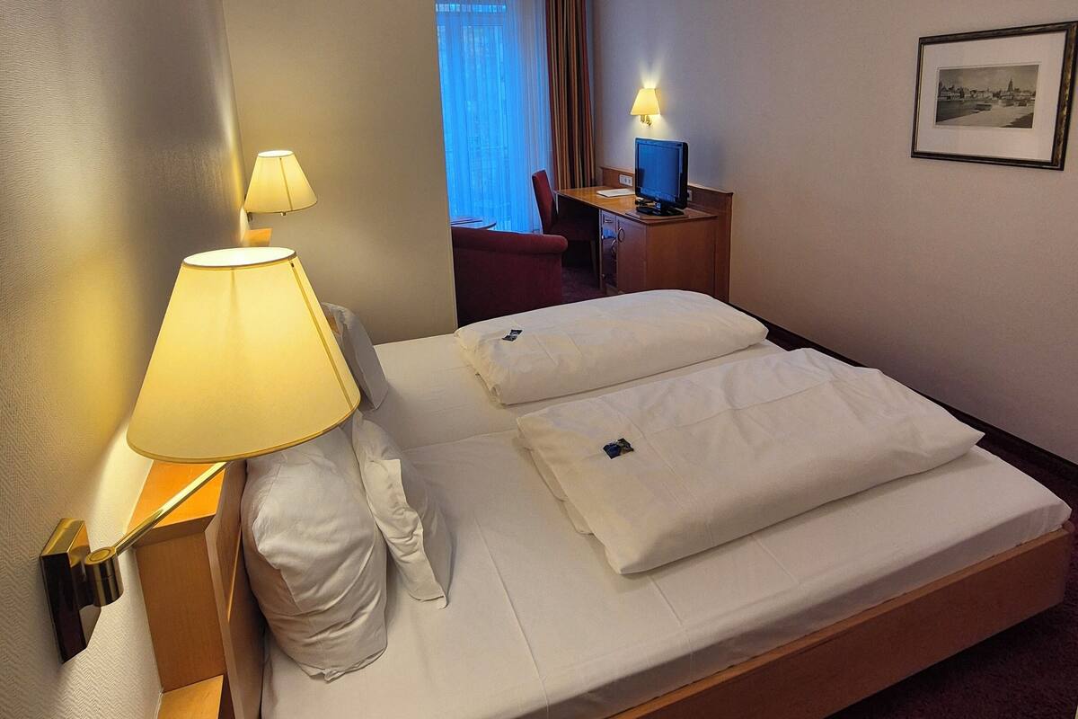 A hotel room with a bed, a desk with a computer, a chair, and a lamp.