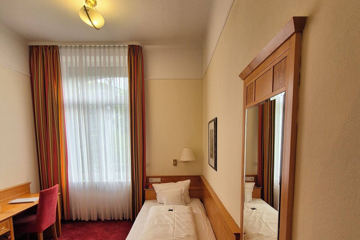 A hotel room with a single bed, a desk, a chair, a mirror, and a window with curtains.