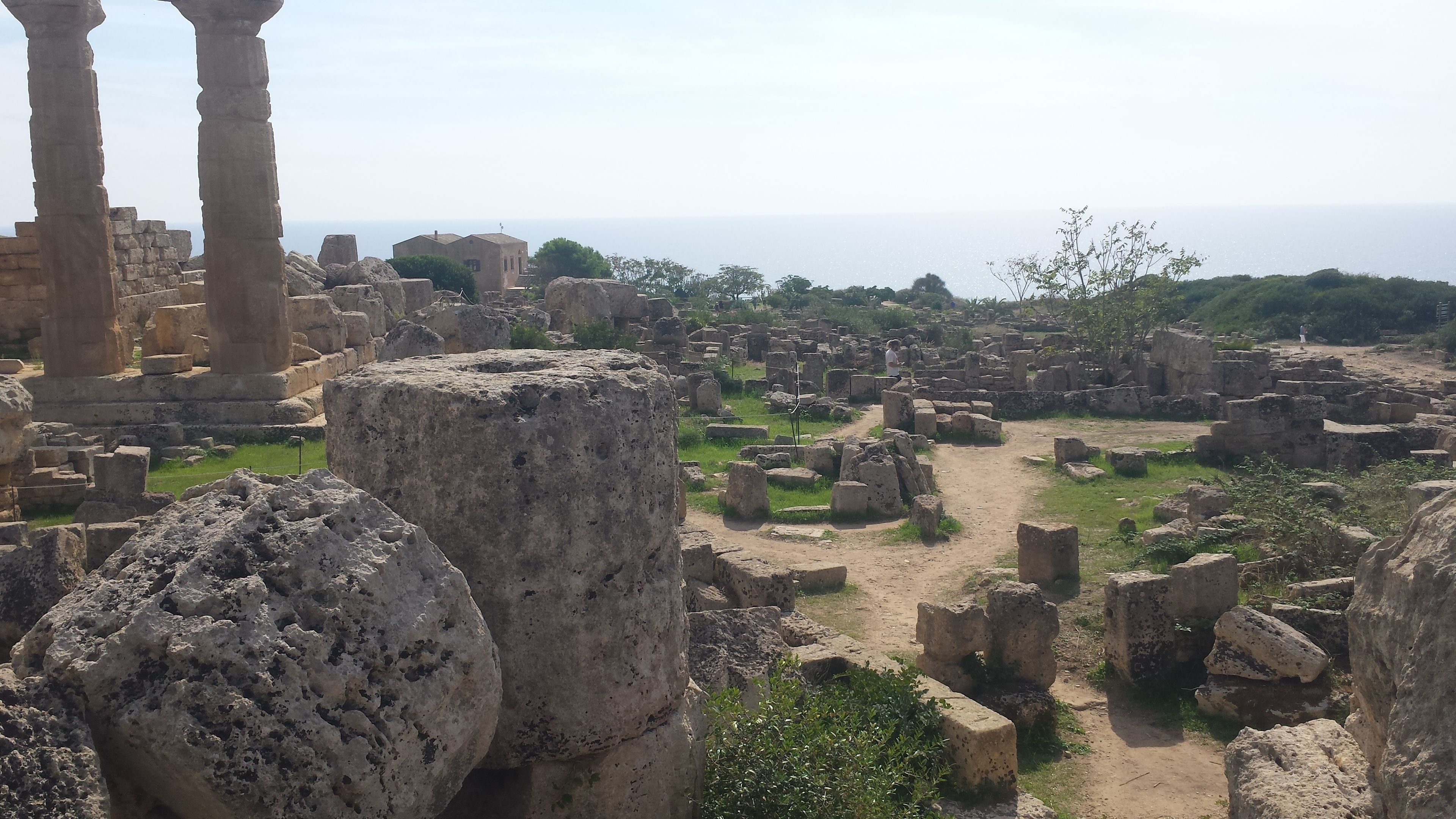 Ancient ruins with stone columns and scattered stone blocks, surrounded by greenery and a clear sky.