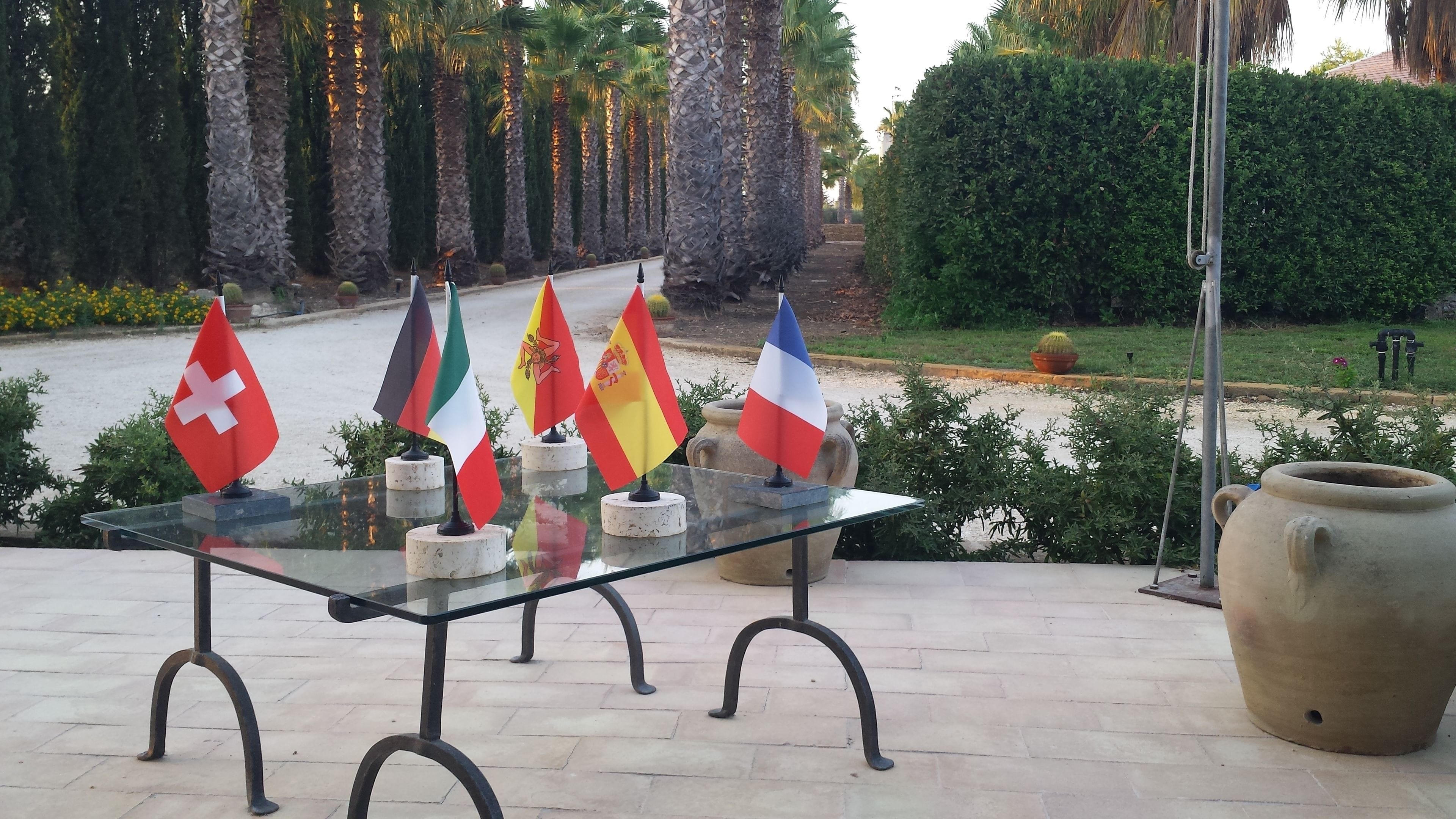 Une table en verre, sur laquelle sont disposés plusieurs drapeaux nationaux sur des supports, entourée d’un jardin planté de palmiers et agrémenté d’un grand pot de fleurs.