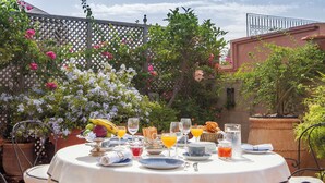 An outdoor dining area with a round table set for breakfast, surrounded by lush greenery and flowering plants. - Dar El Mudal - Adults Only (Marrakech)