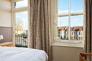 Habitación de hotel con cama, mesita de noche, ventana con cortinas y vista a edificios.