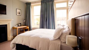 A neatly arranged bedroom with a bed, a bedside table with a lamp, a fireplace, and a window with curtains. - The Wellington (Bristol)