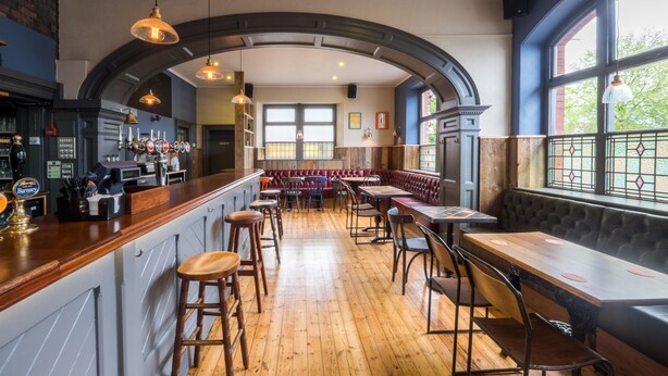 A bar area with wooden flooring, bar stools, and a counter with a variety of bottles. There are tables and benches for seating, and large windows with a view of the outside. - The Wellington (Bristol)