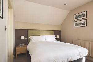 A neatly made bed with white linens, a wooden nightstand with a lamp, and two framed pictures on the wall.