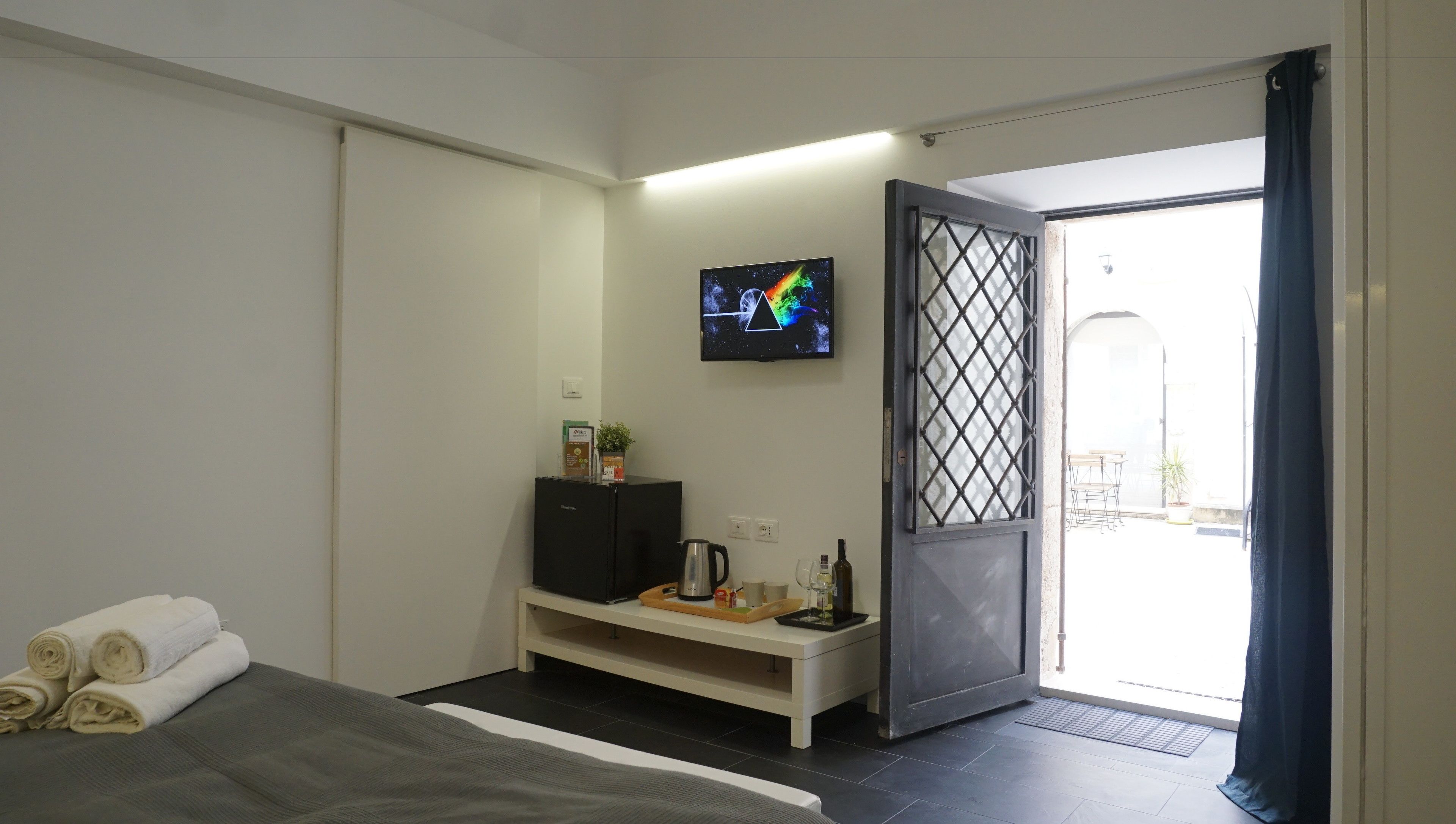 A modern hotel room with a bed, a TV, a small kitchenette, and a sliding door to the balcony.