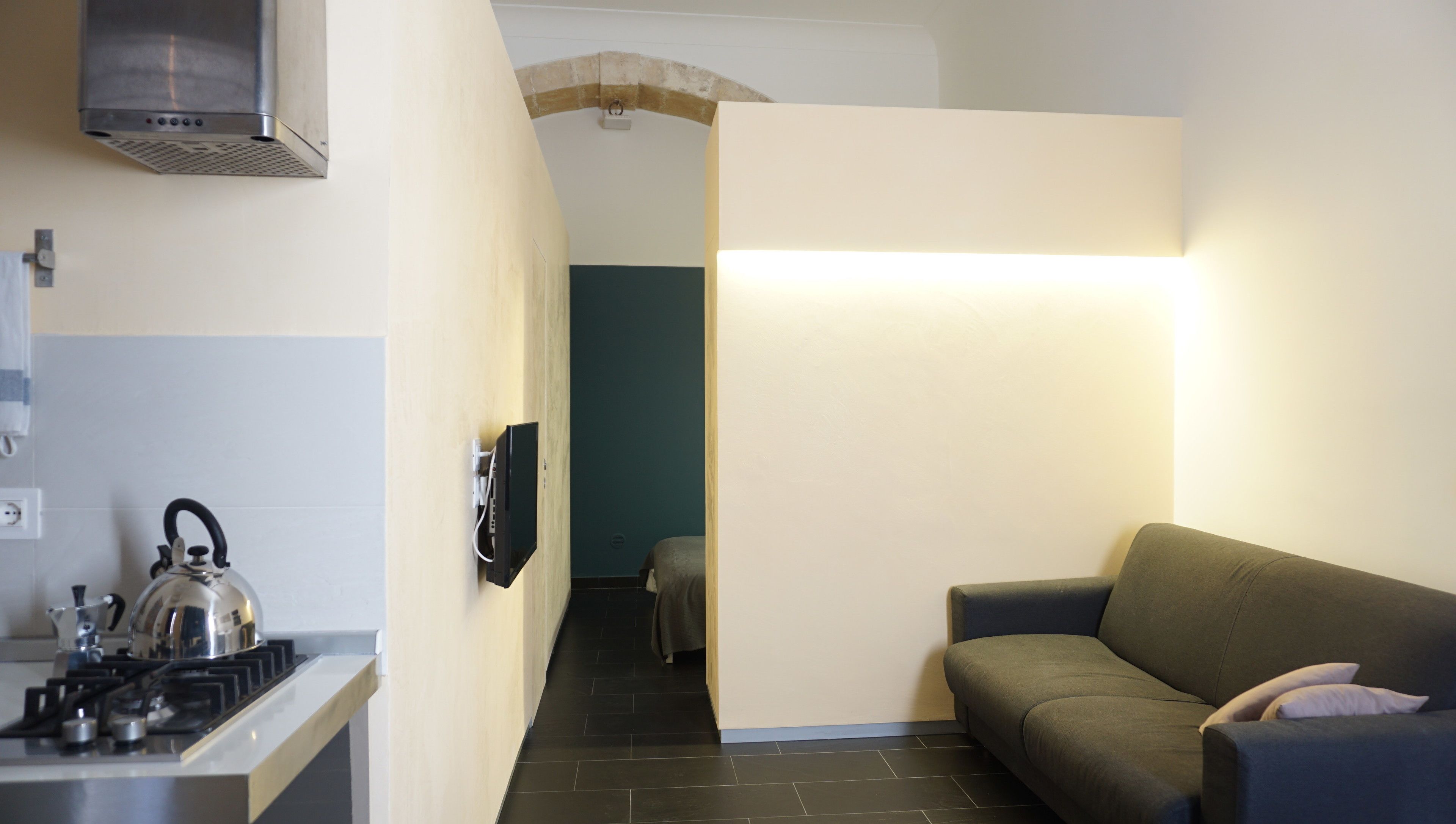 A compact kitchen with a gas stove, kettle, and a TV mounted on the wall. A sofa is placed in the living area, and a hallway leads to another room.