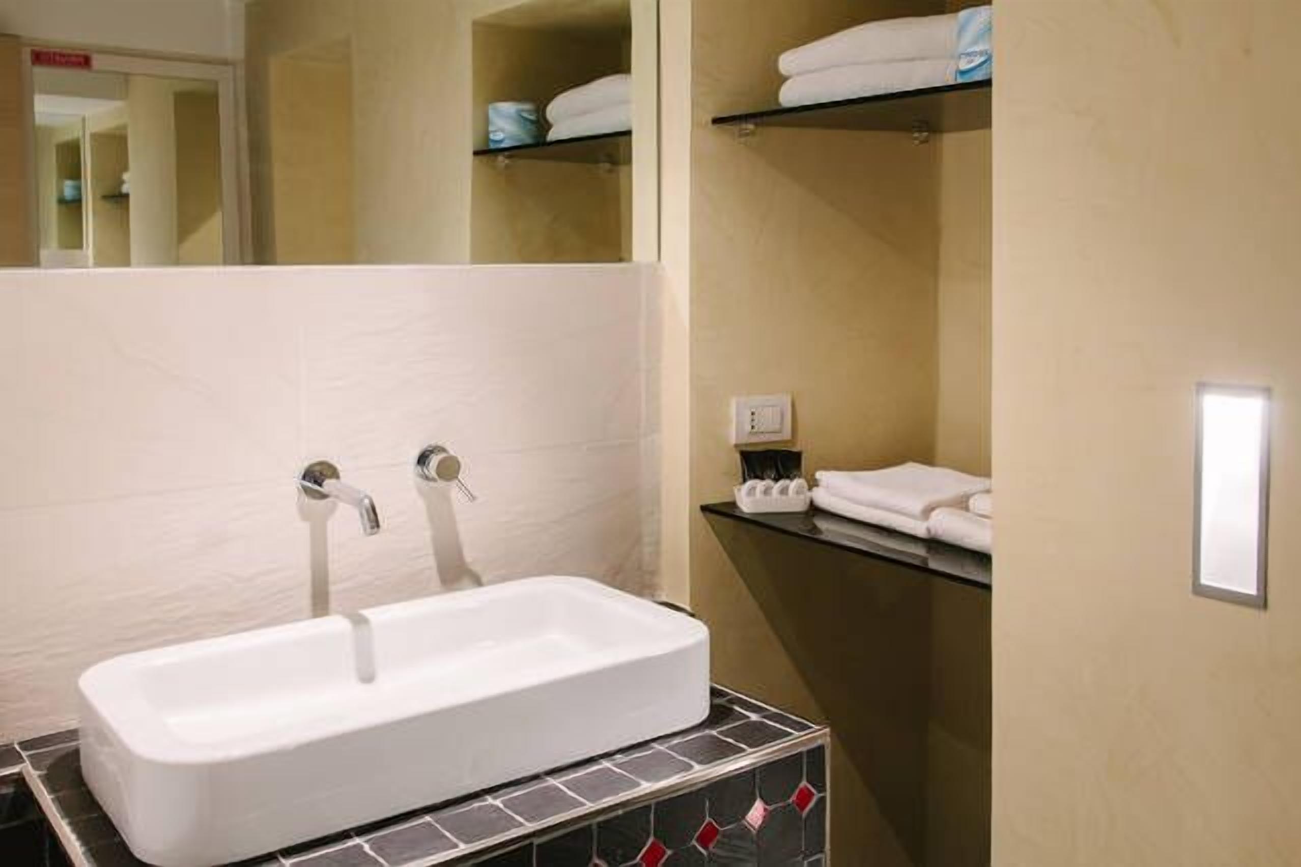 A bathroom with a white sink, two faucets, and a mirror. There are shelves with towels and toiletries.