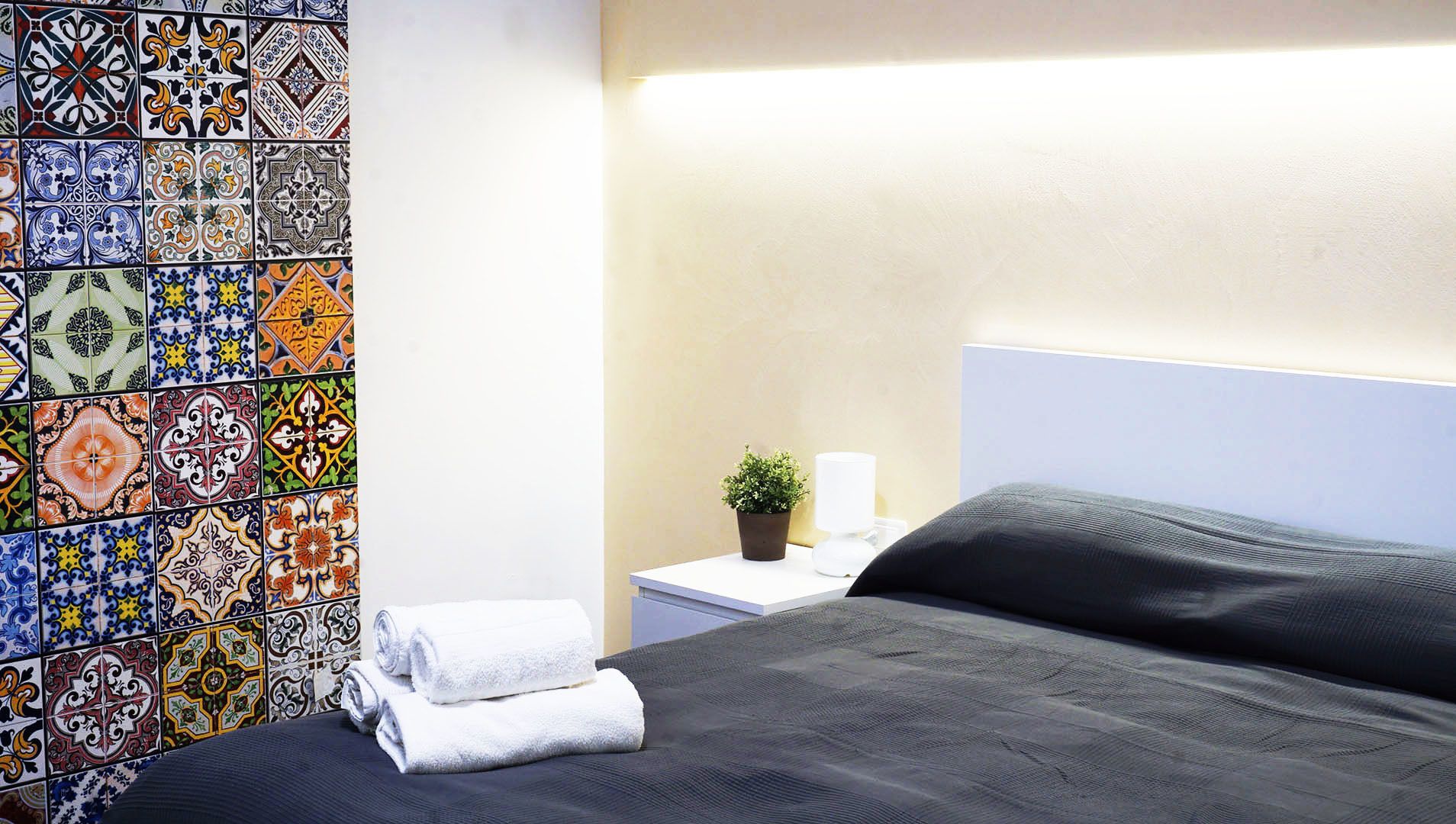 Habitación de hotel con cama, mesita de noche, planta en maceta y pared con azulejos de colores.