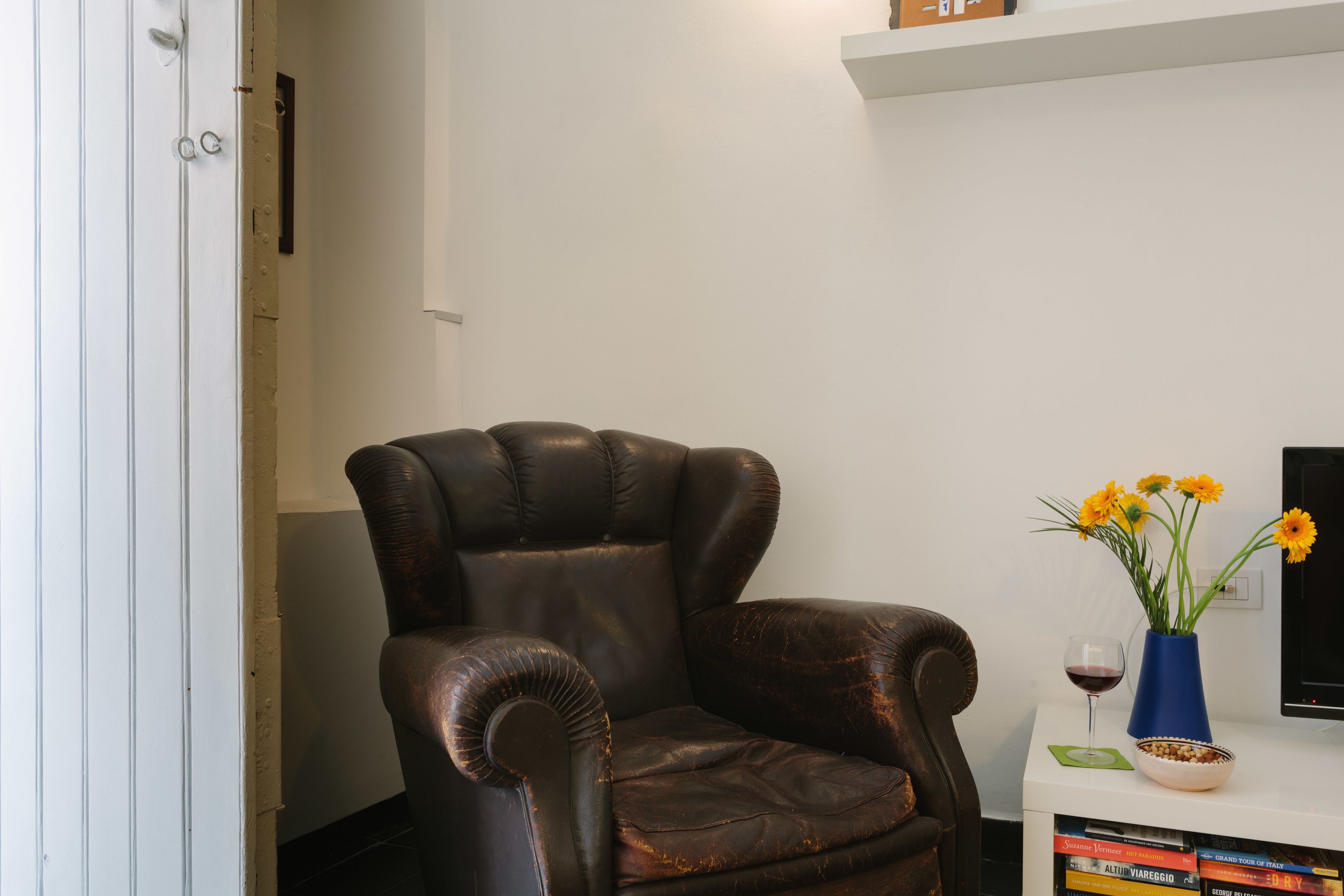 A leather armchair, a vase with flowers, a wine glass, and a stack of books on a white table.