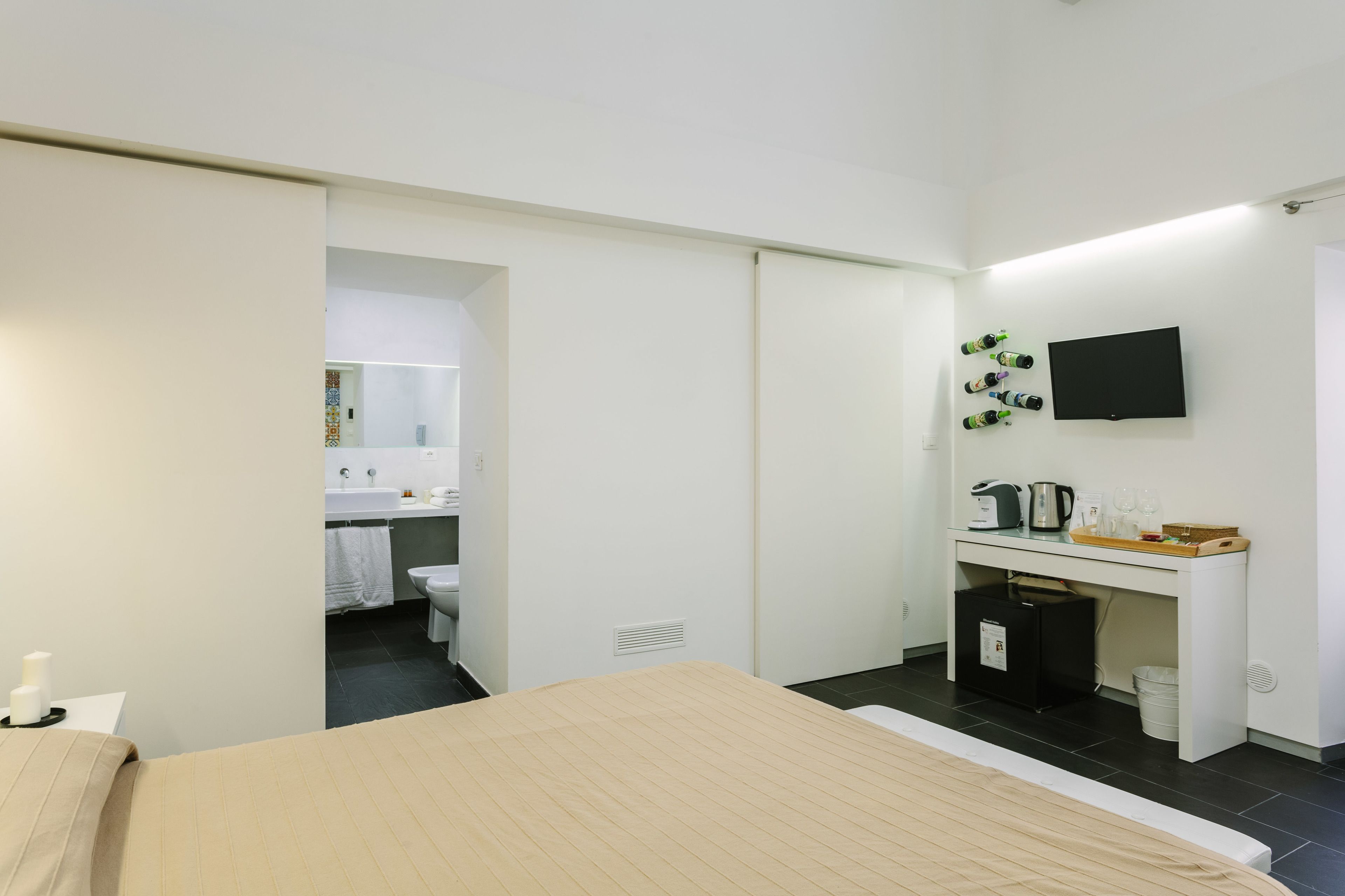 A modern hotel room with a bed, a desk with a flat-screen TV, a small kitchenette, and a bathroom visible through an open door.