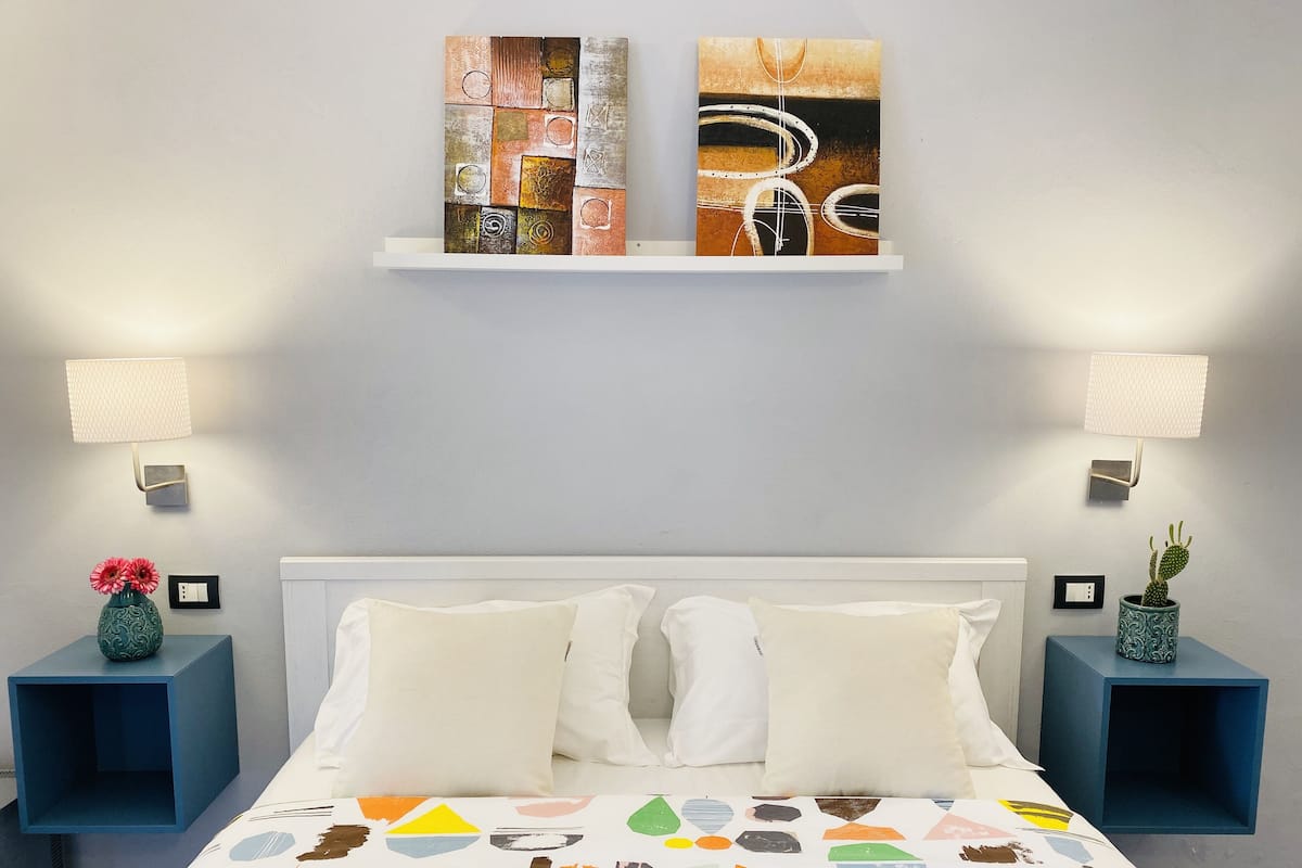 Un dormitorio con una cama, dos mesitas de noche, un estante en la pared con una obra de arte y un cactus sobre una de las mesitas.