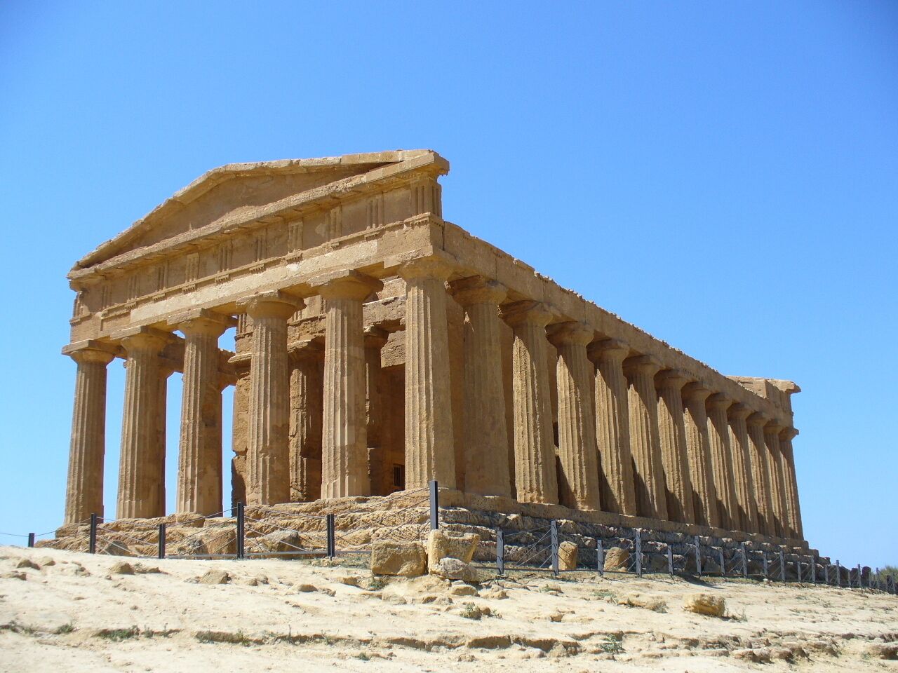 A large ancient Greek temple with multiple columns and a flat roof, situated on a sandy hill.